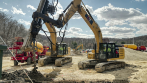 CNC Foundations installing rigid inclusions and vibratory stone columns at a commercial job site using heavy-duty drilling rigs and grout equipment. Yellow excavators with CNC logos operate side-by-side under clear skies, demonstrating ground improvement techniques for soil stabilization and deep foundation support.