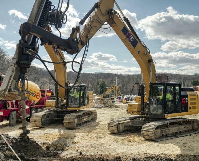 CNC Foundations installing rigid inclusions and vibratory stone columns at a commercial job site using heavy-duty drilling rigs and grout equipment. Yellow excavators with CNC logos operate side-by-side under clear skies, demonstrating ground improvement techniques for soil stabilization and deep foundation support.