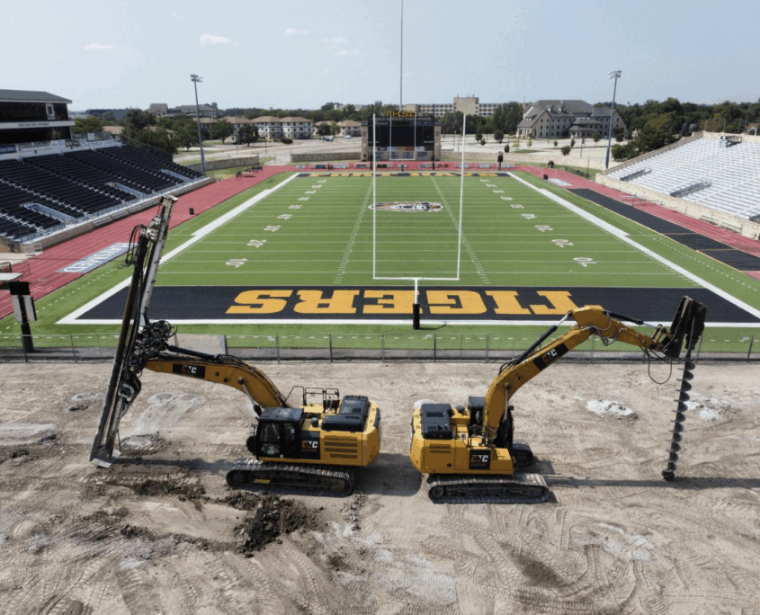 Two CNC Foundations drilling rigs installing aggregate piers in front of a university football field in Hays, Kansas. The image shows the rigs positioned along the end zone marked “TIGERS,” with the stadium and bleachers in the background. This ground improvement work is part of a geotechnical project to support athletic facility upgrades.