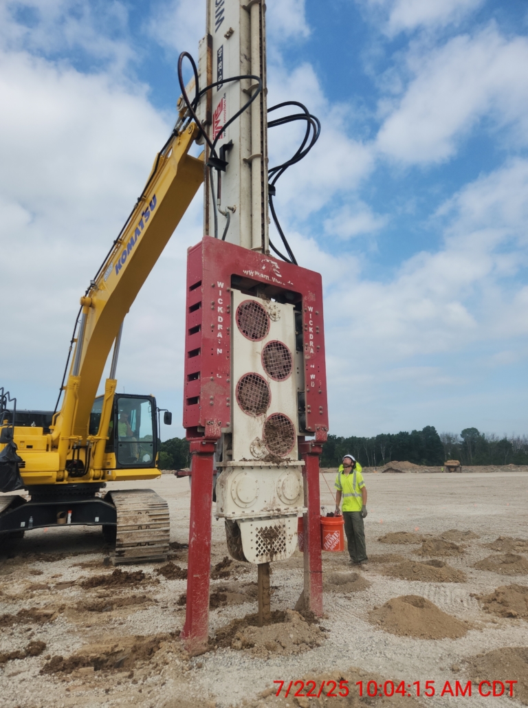 Close-up view of a CNC Foundations operator standing next to a Komatsu excavator-mounted vibratory rig installing wick drains at the Appleton, WI jobsite. Used for soil consolidation and foundation support of warehouse structures.