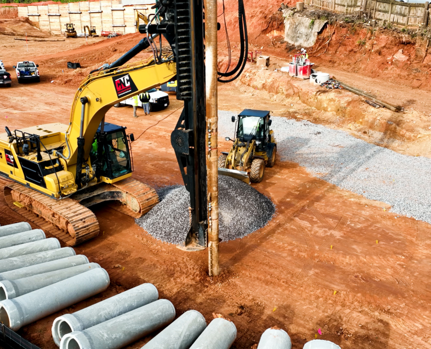 A CNC Foundations excavator installs an aggregate pier using a vertical drill rig on a red clay construction site in Athens, Georgia. A gravel pile sits at the base of the rig, with another CNC loader in the background and rows of concrete casing pipes stacked on the left. Several service vehicles and construction workers are visible near the excavation wall.