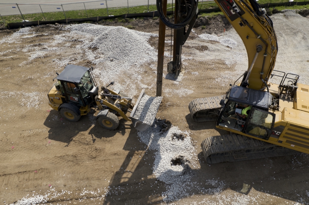 Overhead view of CNC Foundations equipment at work: a yellow front loader is seen delivering crushed stone into a borehole while a large drilling rig installs aggregate piers on a dirt construction site. White crushed rock surrounds the area, indicating active ground improvement in progress for The Row Housing Development in Indianapolis.