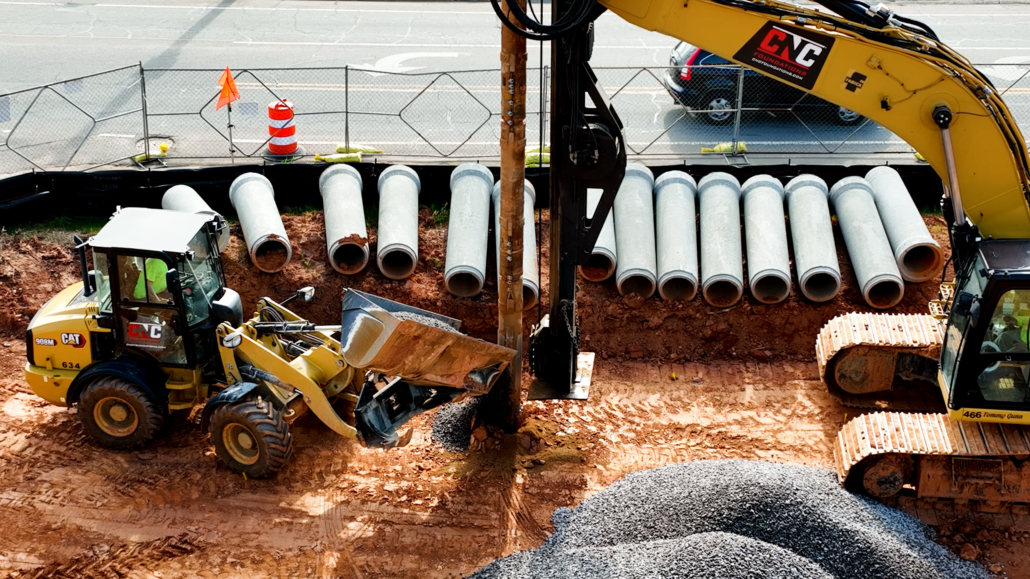 Close-up view of CNC Foundations equipment installing aggregate piers at a university housing site. A loader pours gravel into a drilled hole while the drill rig holds steady. Behind the equipment, large concrete pipes are lined along a safety fence near a roadway, and a CNC-branded excavator is positioned for support.