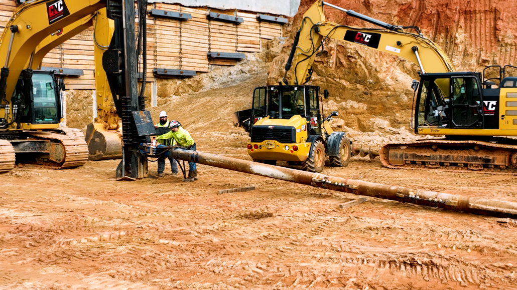 CNC Foundations crew members prepare a drill shaft for installation of aggregate piers on a compacted red clay site. Two workers in PPE guide the drill while surrounded by heavy machinery, including CNC-branded excavators and loaders. A soldier pile wall is visible in the background, supporting the excavation zone.