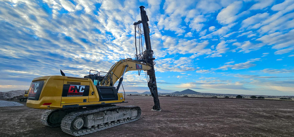 A CNC Foundations rig equipped with a vibratory compaction tool stands under a dramatic cloud-filled sky at SunZia Wind Farm during aggregate pier ground improvement for wind turbine infrastructure.