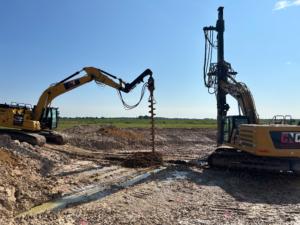 crossover wind_cnc foundations_Settlement Control Solutions2 Two CNC Foundations drilling rigs in operation on a muddy construction site, with one auger actively drilling into the ground to install foundation support for wind turbines under clear blue skies.