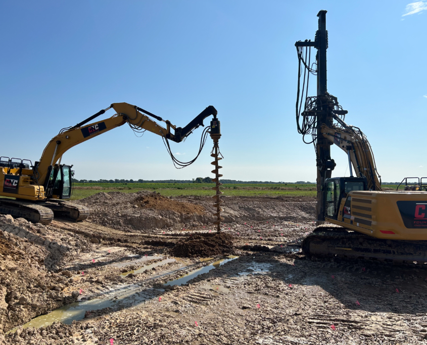 Two CNC Foundations drilling rigs in operation on a muddy construction site, with one auger actively drilling into the ground to install foundation support for wind turbines under clear blue skies.