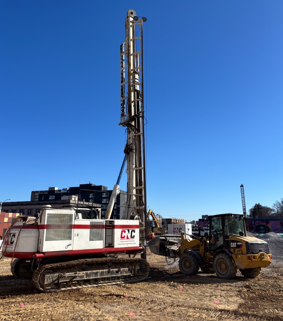 delmar loop_cnc foundations_AGGREGATE PIER INSTALLATION2 Close-up of a CNC Foundations drill rig on tracked treads alongside a CAT 908M wheel loader at the Delmar Loop site. The equipment is positioned on cleared soil with mid-rise buildings in the background and a deep blue sky overhead.