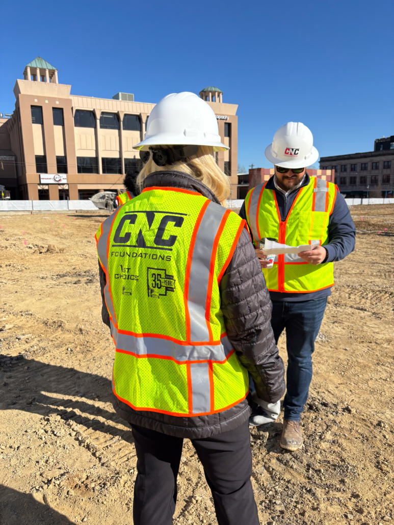 delmar loop_cnc foundations_AGGREGATE PIER INSTALLATION3 Two CNC Foundations team members reviewing plans on site in front of a casino building. Both are wearing CNC-branded safety vests and white hard hats, with the team member in the foreground displaying a vest commemorating CNC’s 35th year in business.
