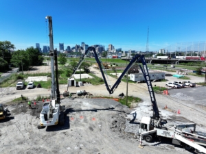 default A wide-angle view of CNC Foundations' rigid inclusion installation at the Riverchase Multifamily Development site in Nashville, TN. A large drilling rig and concrete pump truck are actively working in tandem under a clear blue sky, with the Nashville skyline visible in the background. Construction crews and vehicles are positioned around the site, indicating active soil stabilization work in progress.