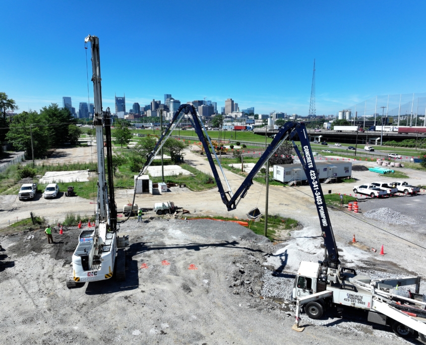 default A wide-angle view of CNC Foundations' rigid inclusion installation at the Riverchase Multifamily Development site in Nashville, TN. A large drilling rig and concrete pump truck are actively working in tandem under a clear blue sky, with the Nashville skyline visible in the background. Construction crews and vehicles are positioned around the site, indicating active soil stabilization work in progress.