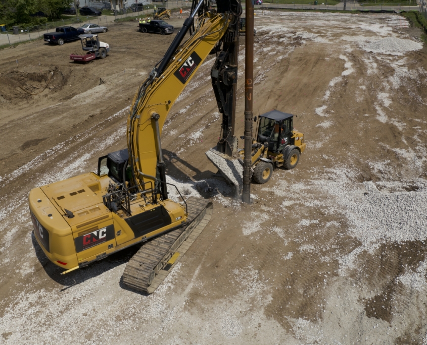 A CNC Foundations aggregate pier drilling rig and a front loader coordinate to place crushed aggregate into a borehole. The jobsite is visibly marked by spread-out white gravel, compacted dirt, and active heavy machinery working to stabilize the ground for residential development in Indianapolis.